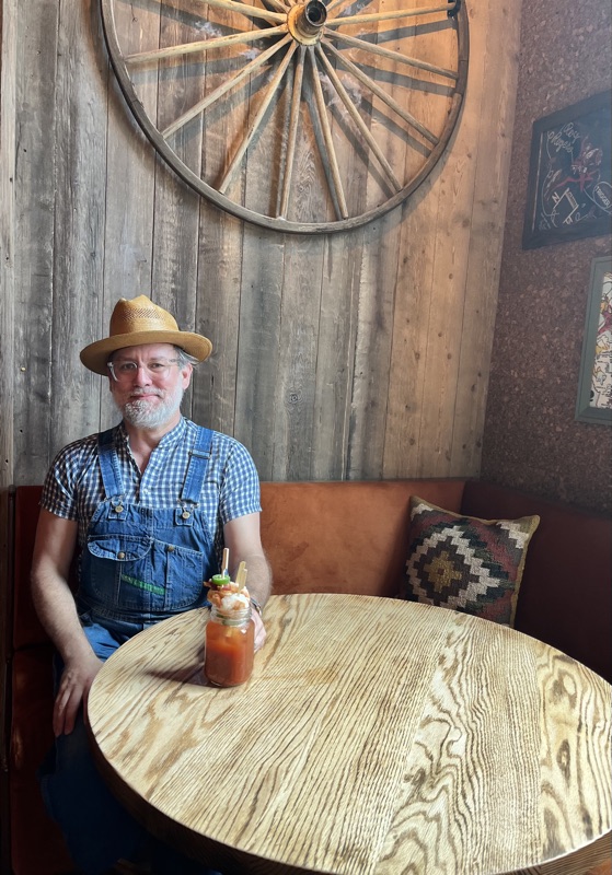 Matt Morelock in a straw hat and overalls, seated at a wooden table beneath a wagon wheel